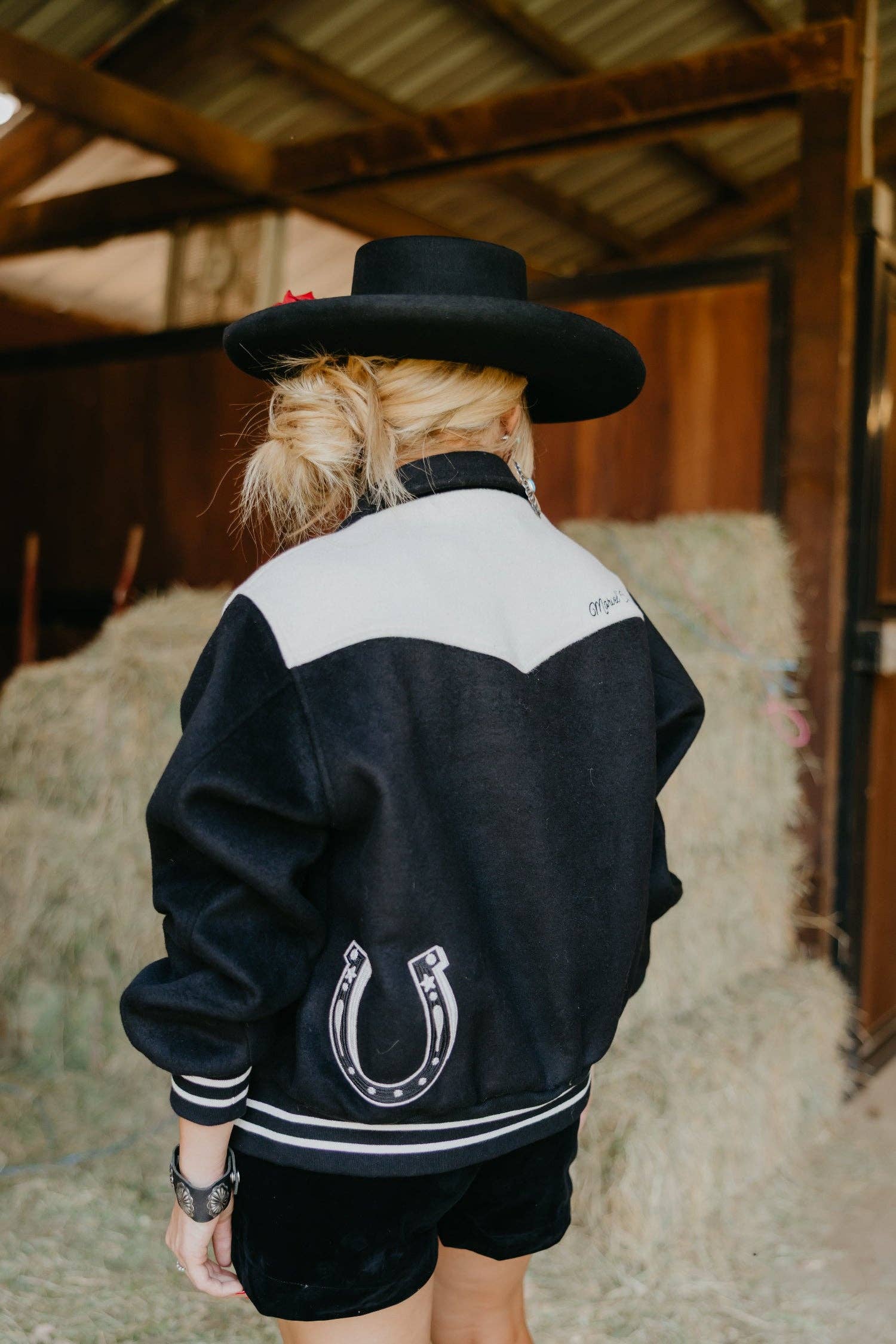 Person wearing a black and white jacket with a horseshoe emblem in a barn setting